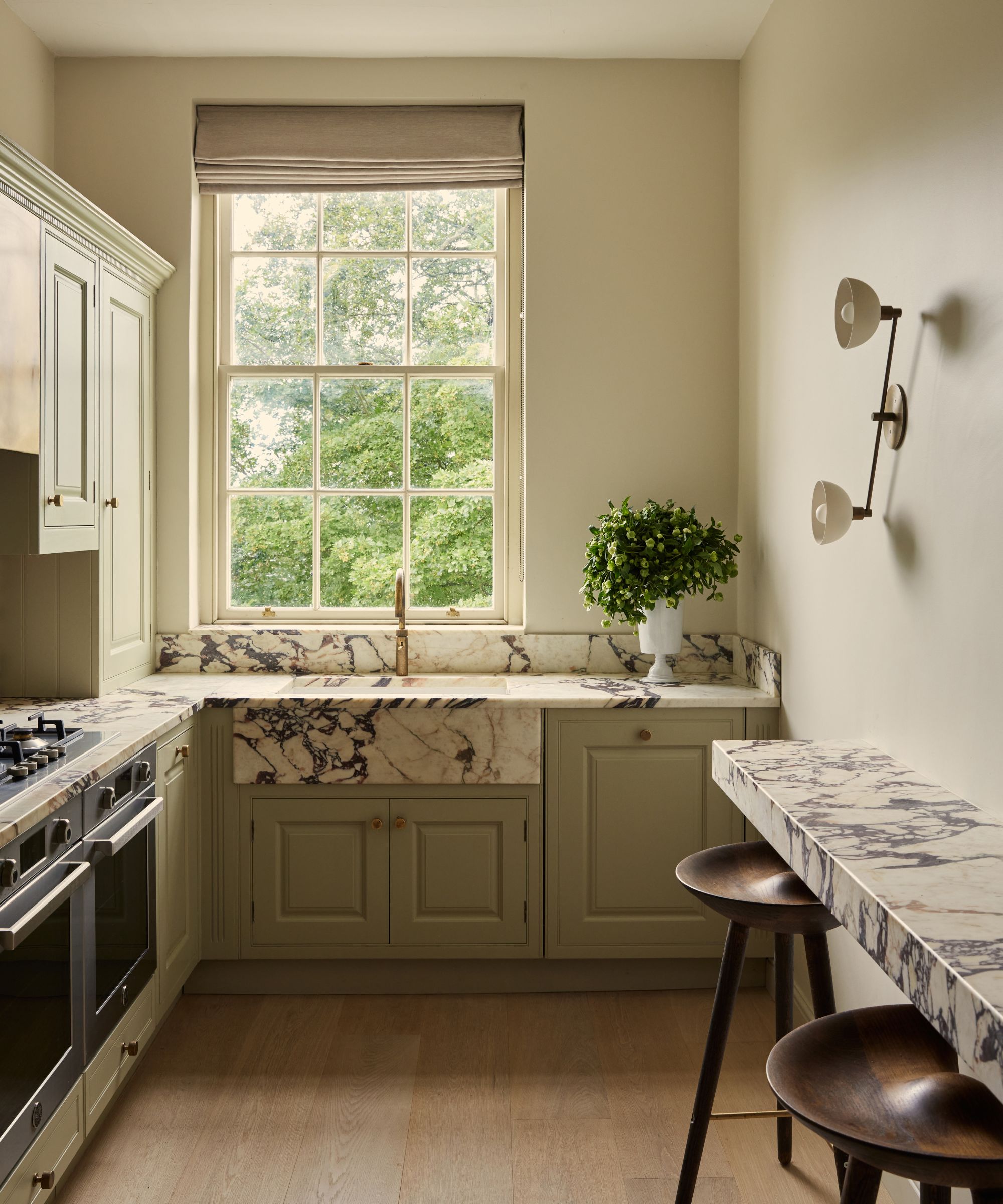Kitchen with large original window, marble sink and backsplash, sage green painted cabinetry with gold knobs, and a breakfast bar built into the side wall with dark mango wood stools