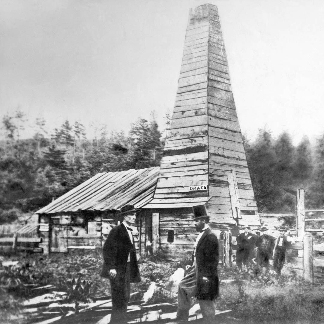 An old black and white photo of Edwin Drake's first oil well. The photo features men in suits and tophats standing in front of the well, and a large wooden structure
