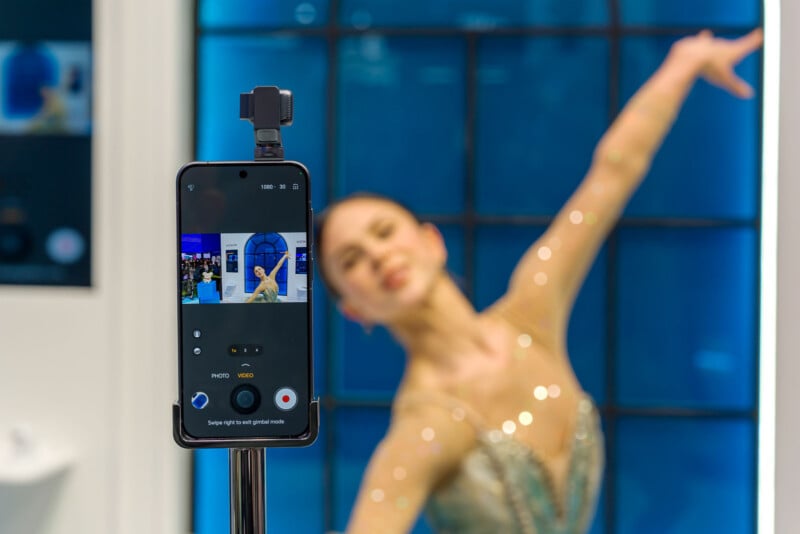 A smartphone on a tripod records a woman in a sparkly dress dancing with one arm raised in front of a large blue window. The phone screen shows her pose in the camera preview.