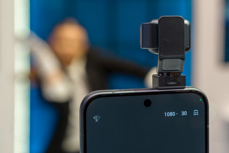 A smartphone with a mounted camera accessory records a video, displaying "1080 30" on the screen. In the blurred background, a person in a suit appears to be posing or dancing in a blue-lit room.