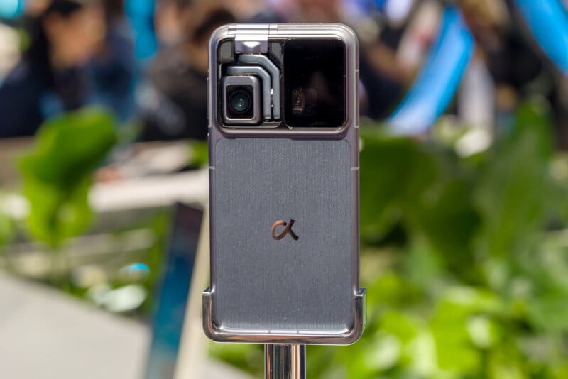 A close-up of a smartphone with a prominent camera module and the Alpha (α) logo on the back, displayed upright against a blurred background with green plants and people.