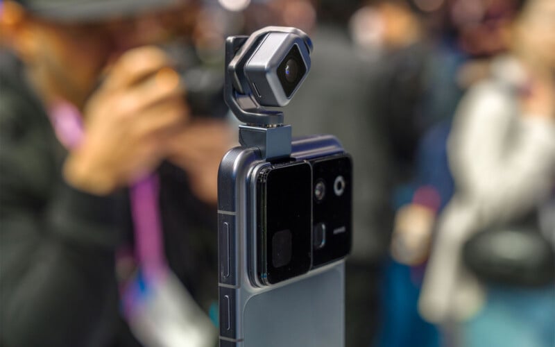 Close-up of a smartphone with a mounted camera accessory at an event, with a blurred person holding a camera in the background.