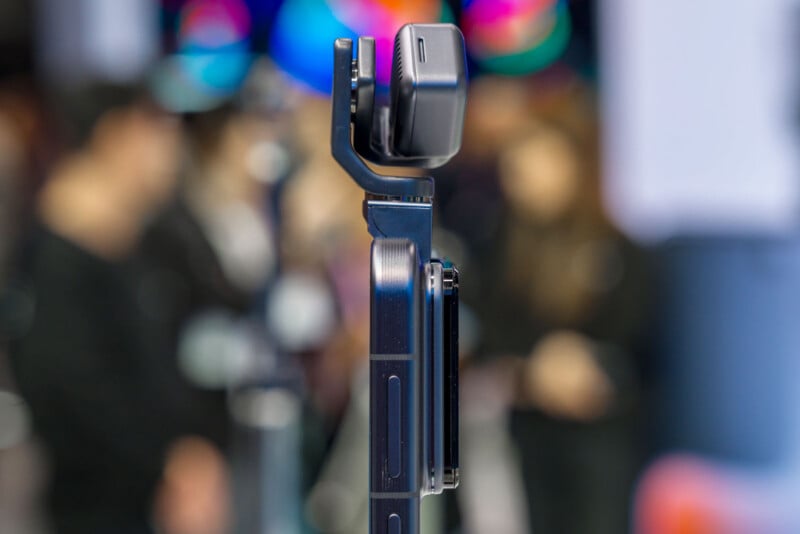 Close-up of a smartphone mounted on a handheld gimbal stabilizer, with a blurred background showing several people in an indoor setting.
