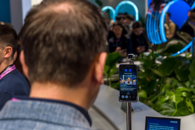 A man stands in front of a smartphone mounted on a tripod, taking a group selfie at a busy tech event with people, greenery, and blue neon lights in the background.