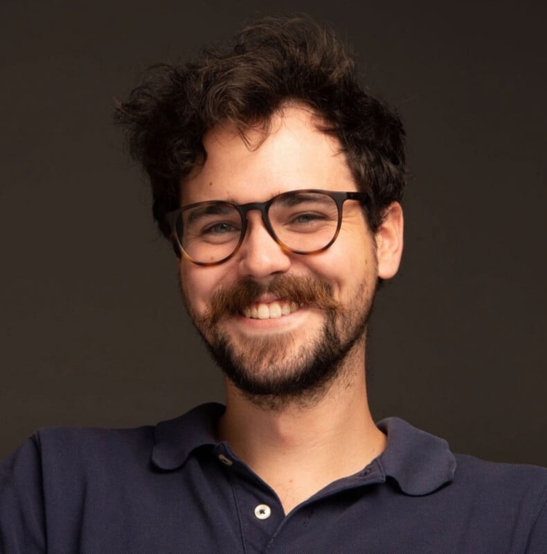 A young man with curly dark hair, a beard, and mustache smiles while wearing black-rimmed glasses and a navy blue polo shirt against a plain dark background.