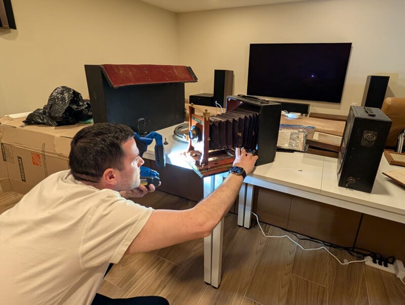 A person examines an antique bellows camera on a table using a flashlight. The table is cluttered with vintage camera cases and other equipment, and cardboard boxes are visible in the background.