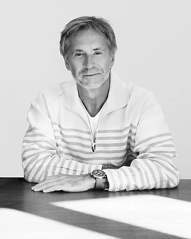 Man with a beard sitting at a table, wearing a striped sweater, looking into the camera in a black and white photo.