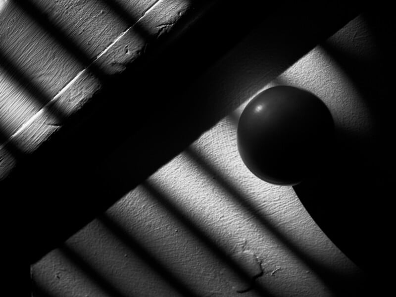 Black and white close-up of a round door knob casting a shadow on a textured door, with diagonal shadows from window blinds creating a dramatic light pattern across the surface.