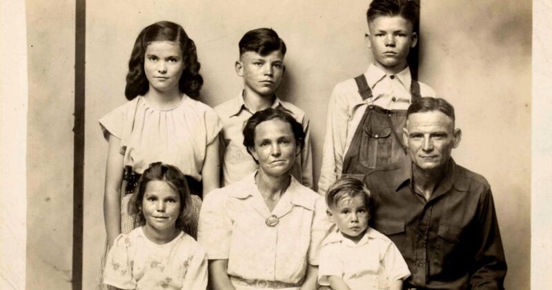 A black-and-white portrait of a family from the early 20th century, showing two adults and four children. The adults are seated in front, with the children standing and sitting around them, all facing the camera with neutral expressions.