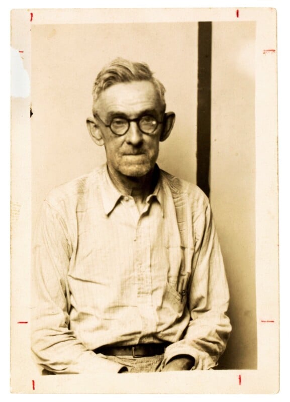 A black-and-white portrait of an older man with glasses, short gray hair, and a mustache, wearing a striped button-up shirt. He is seated against a plain background, looking slightly past the camera.