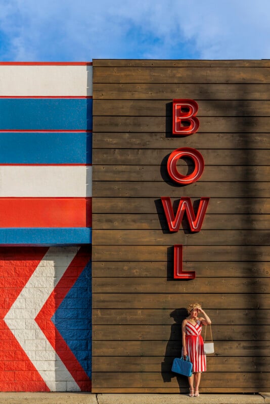 A woman in a red and white striped dress stands in sunlight next to a brown wall with large red letters spelling "BOWL." The wall is adjacent to a building painted with red, white, and blue stripes and a large arrow.