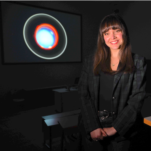 Smiling woman with long hair standing in a classroom. An image of Uranus with its rings is on the wall behind her.