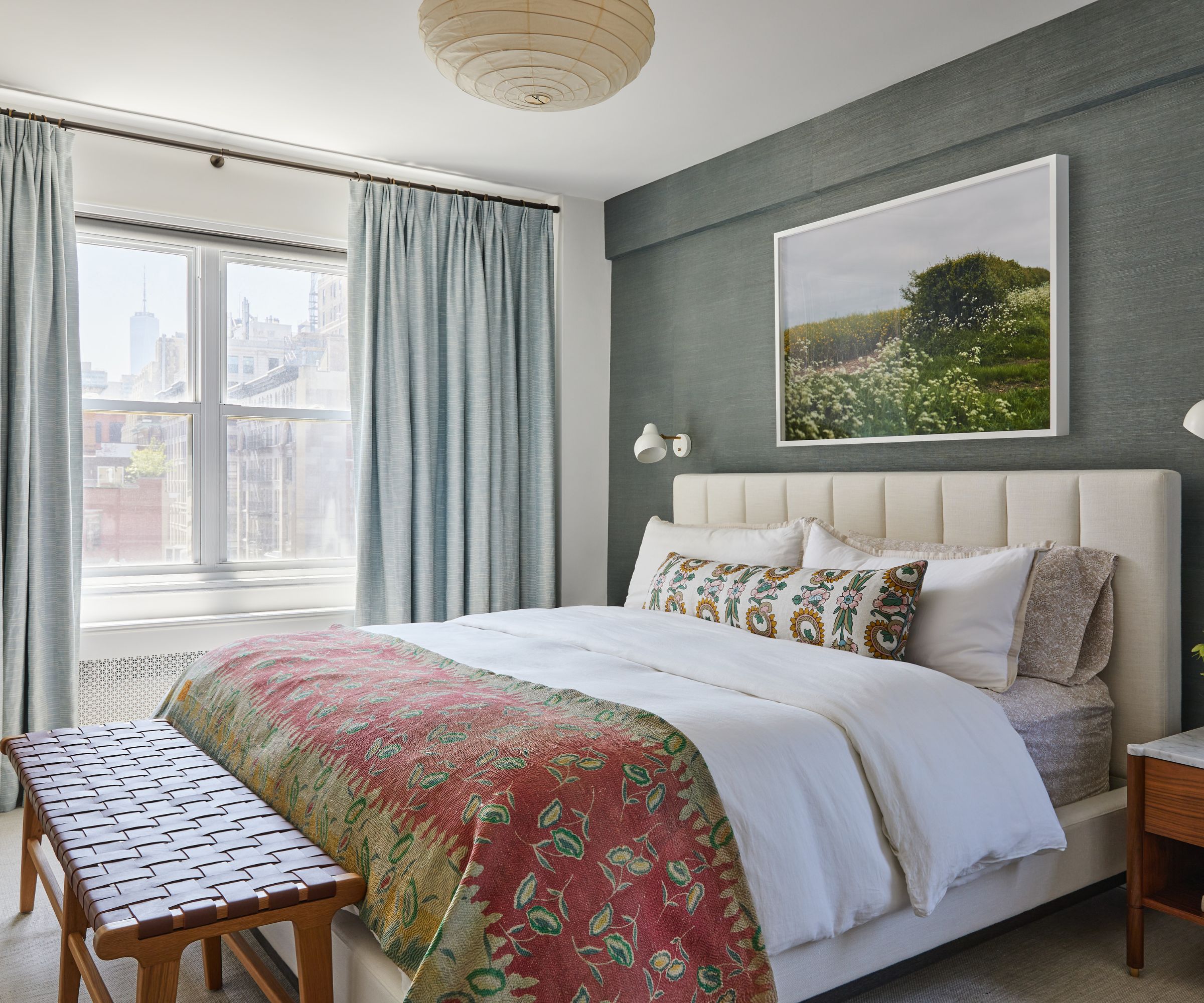 A bedroom with a gray wallpapered wall, light blue curtains, a large cream-colored headboard with white bedding, and a pink patterned throw. A wooden bench at the end of the bed with a leather top.