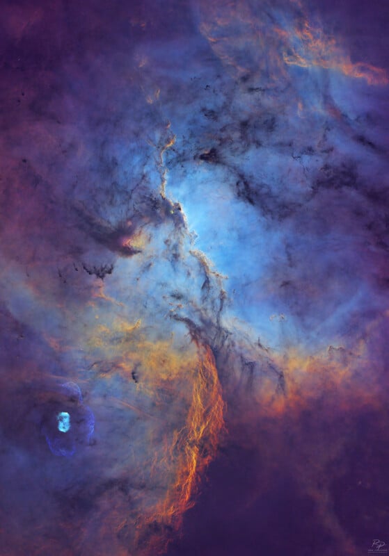 Bright nebula with swirling blue and purple clouds, accented by orange and yellow streaks; cosmic dust and glowing gas create a dramatic celestial scene against a dark background.