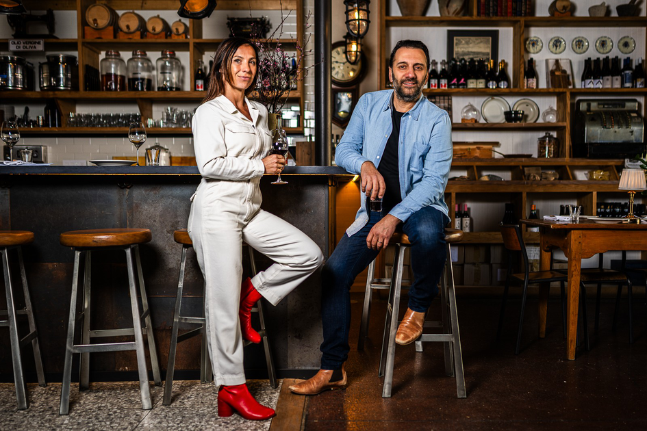 A woman in a white jumpsuit and red boots stands with a wine glass, while a man in a blue shirt and jeans sits on a bar stool holding a glass.