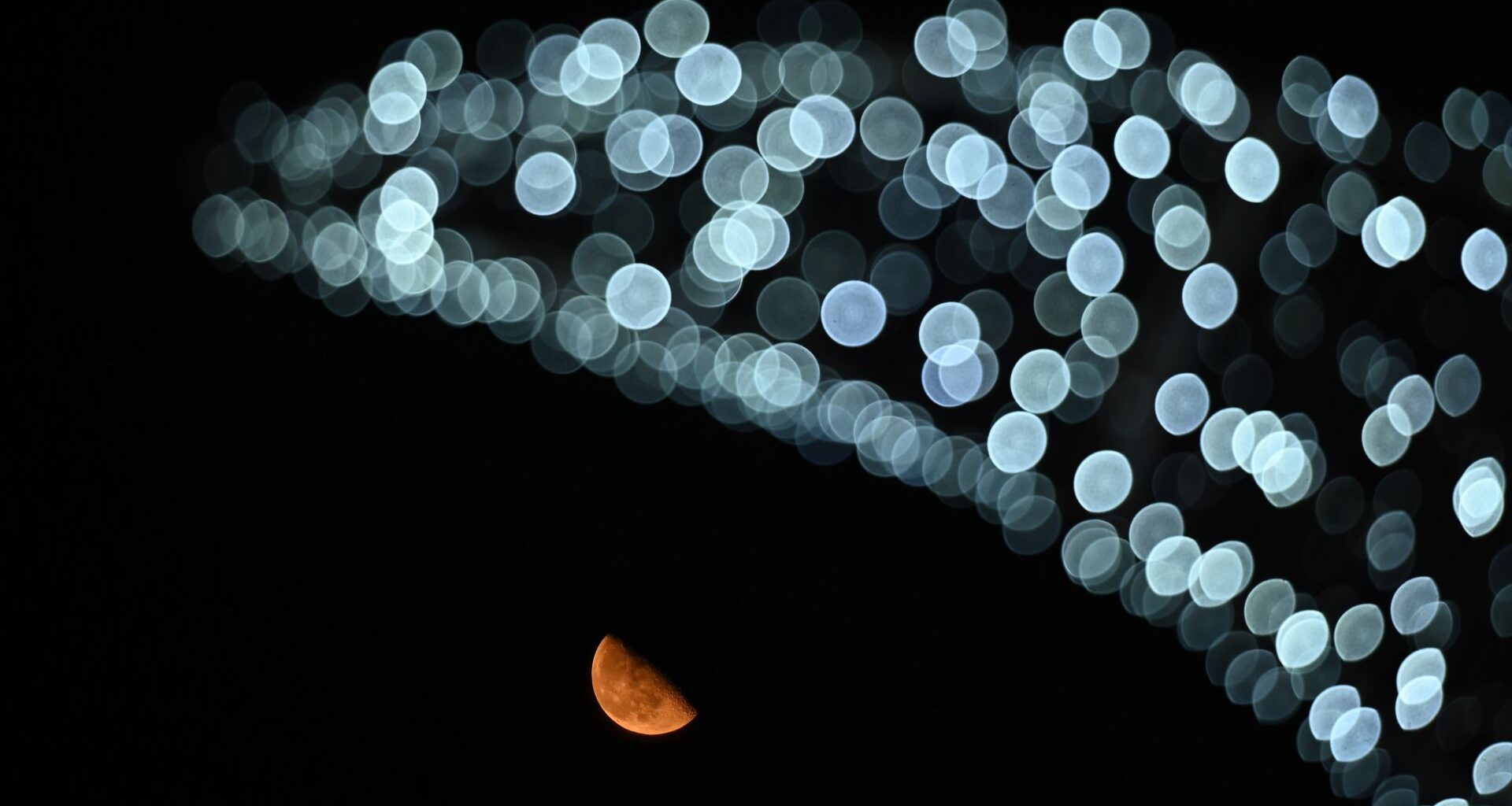 A half-lit moon shines beneath out-of-focus artificial lights arrayed in an arc against a black sky.