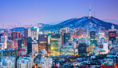 A vibrant cityscape of Seoul at dusk, with brightly lit skyscrapers in the foreground and Namsan Mountain topped by the illuminated N Seoul Tower in the background. The sky transitions from blue to purple.