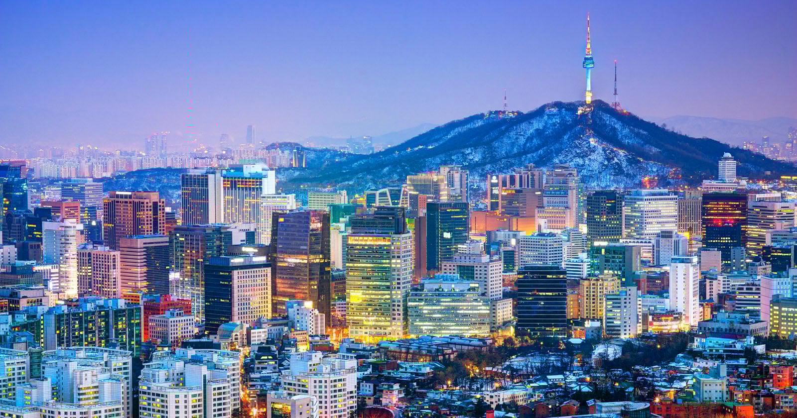 A vibrant cityscape of Seoul at dusk, with brightly lit skyscrapers in the foreground and Namsan Mountain topped by the illuminated N Seoul Tower in the background. The sky transitions from blue to purple.