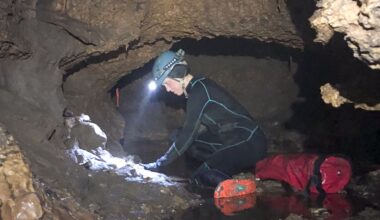 A person wearing a black suit and hard hat with a flashlight on it bends over a circular wall of a small cave.