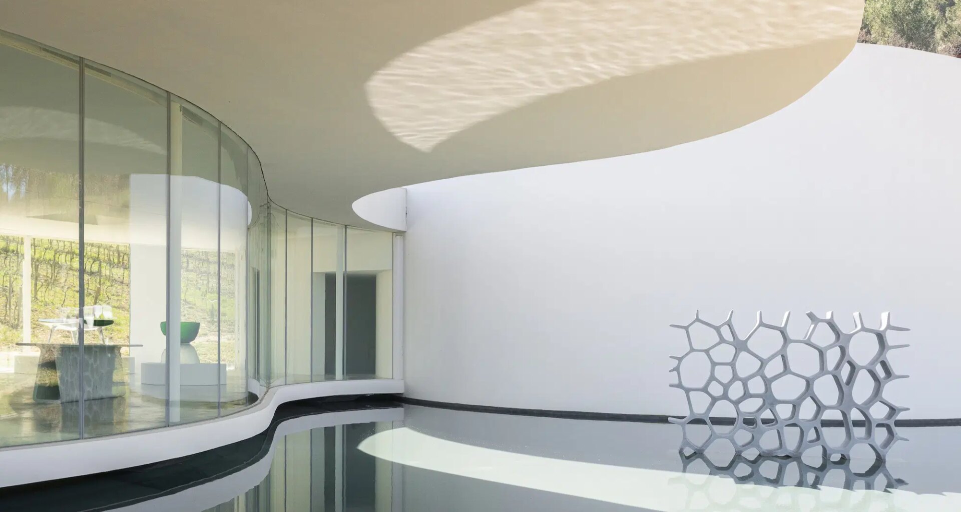 Modern architectural design with curved glass walls and a honeycomb-like sculpture by a reflective water surface.