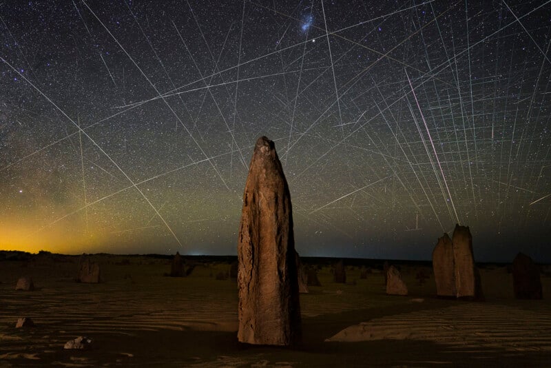Tall limestone rocks rise from a sandy desert under a clear night sky filled with stars and intersecting bright streaks of light, possibly meteors or satellites, creating a dramatic celestial pattern.