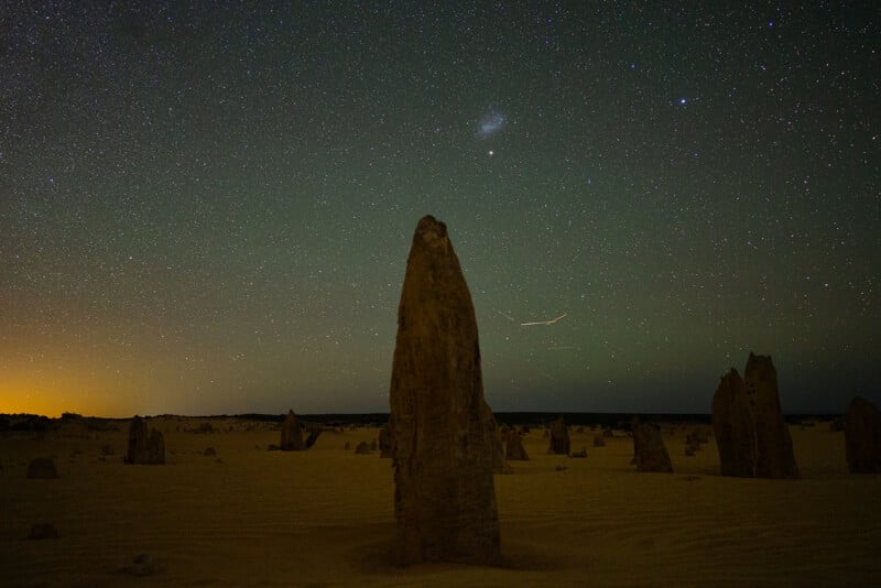A tall, jagged limestone rock stands in a sandy desert under a clear, star-filled night sky with faint light on the horizon. Smaller rock formations are scattered throughout the scene.