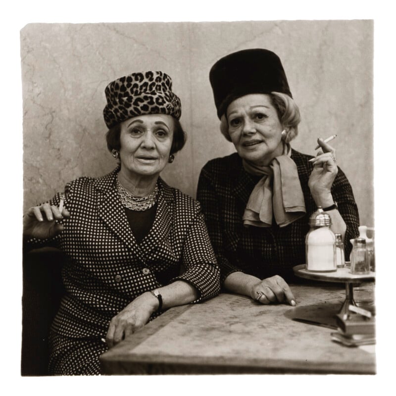 Two older women sit at a table, both wearing hats and patterned outfits. One woman holds a cigarette, and there are small bottles on the table. They look at the camera with neutral expressions. The photo is in black and white.