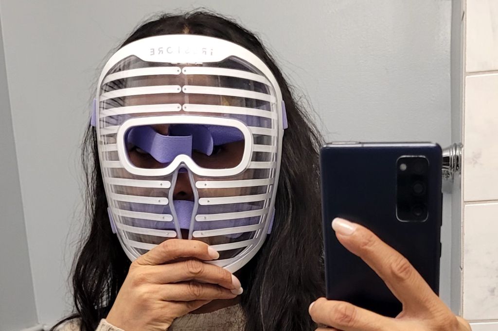 A person with long dark hair wearing an iRestore mask over their face and holding up a smartphone to take a photo.