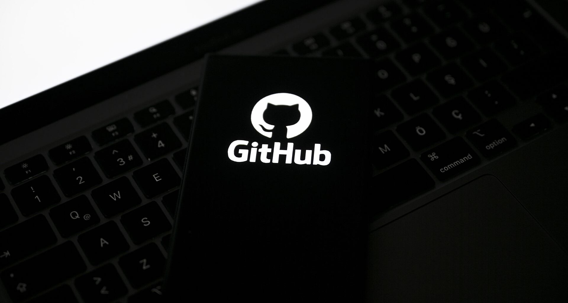 GitHub logo pictured on a smartphone placed on top of a laptop keyboard, with blank white screen in corner.