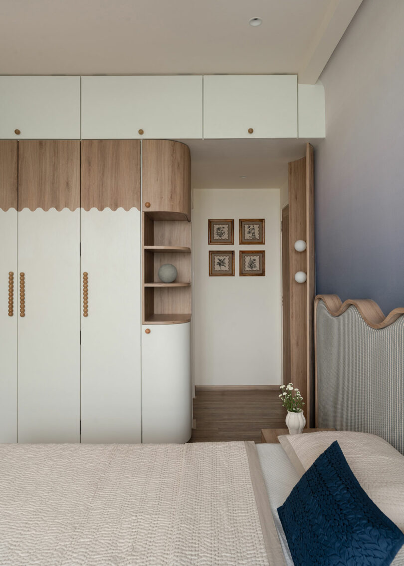 Modern bedroom with built-in white and wood wardrobes, a beige bed with blue pillow, and three framed pictures on the hallway wall.