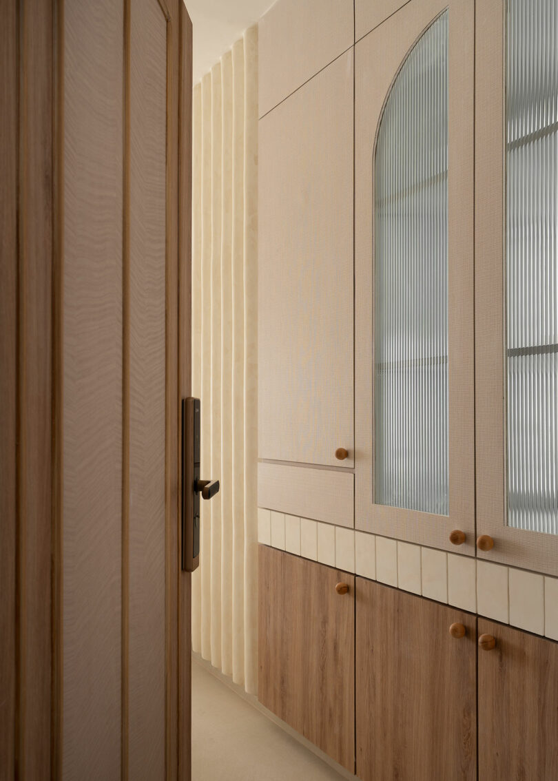 A partially open wooden door reveals built-in cabinets with glass and wood paneling, round knobs, and vertical wall detailing in a minimal, neutral-toned room.