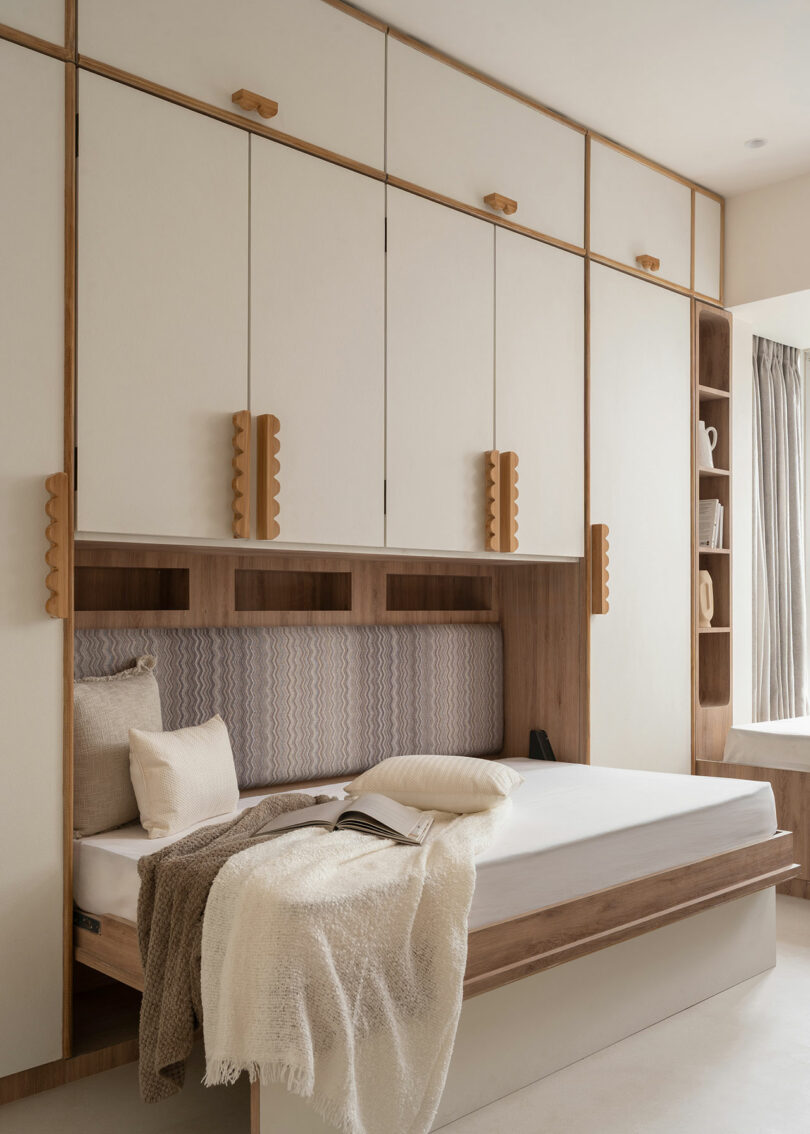 Minimalist bedroom with a wood-framed bed, neutral bedding, built-in cabinets above, and shelves on the right; natural light enters through a window with beige curtains.