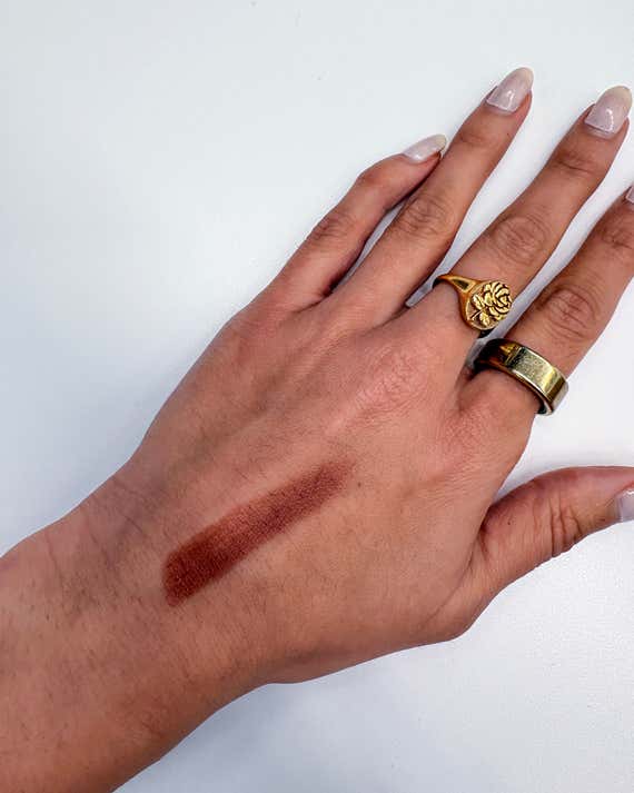A hand with gold rings and long nails rests against a white surface, featuring a brown swatch of Haus Labs Color Fuse Glassy Blush Balm Stick on the back of the hand.