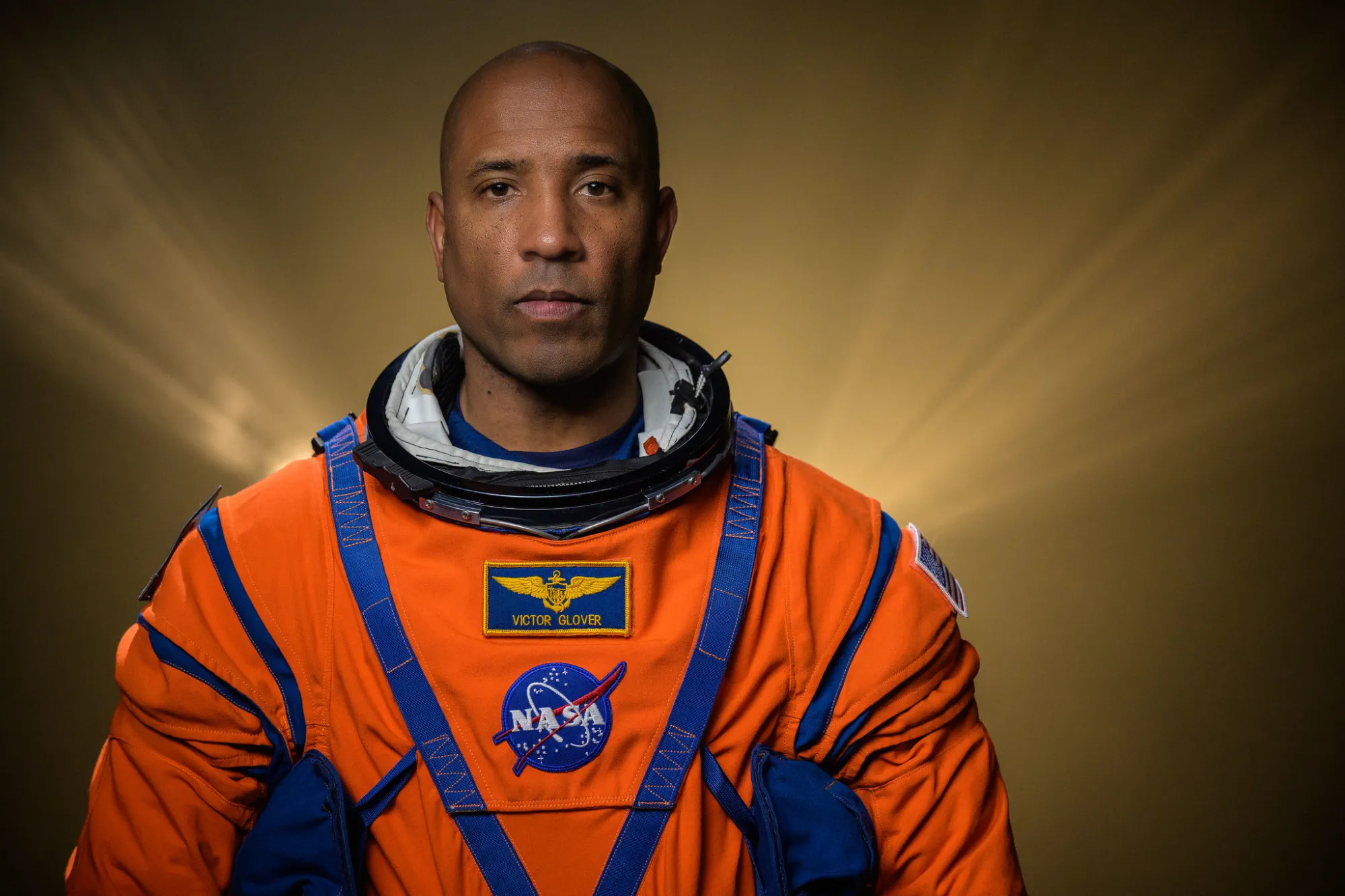 An African American man wearing an orange spacesuit looks at the camera with lights streaking softly up behind him with a tan wall in the background