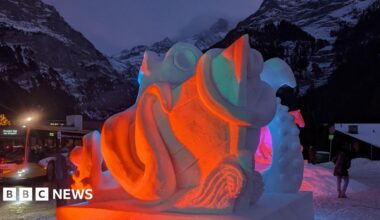 Red and pink lights on a snow sculpture of a sea monster and a ship with snow covered mountains and a bus in the background