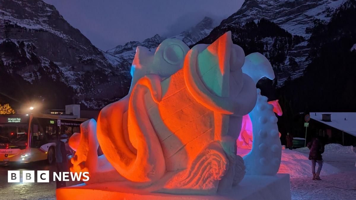 Red and pink lights on a snow sculpture of a sea monster and a ship with snow covered mountains and a bus in the background