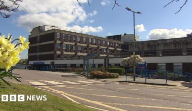 The outside of a hospital on a clear sunny day. The building has lots of windows and has the usual NHS branding on the front of it. There is a road in front of it with some flowers to the left of the pictures.