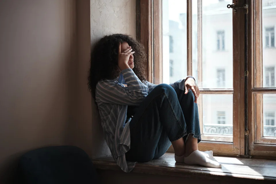 A person sitting on a windowsill, covering their face with their hand, and looking out at a cityscape