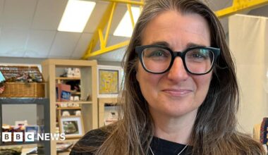 A head and shoulders image of Melissa Santiago-Val. She is wearing a black top and a necklace and glasses. She is looking directly into the camera. Behind her is some shelving with various items on it and to her left is a headless mannequin with handmade jewellery and clothing hanging on it.