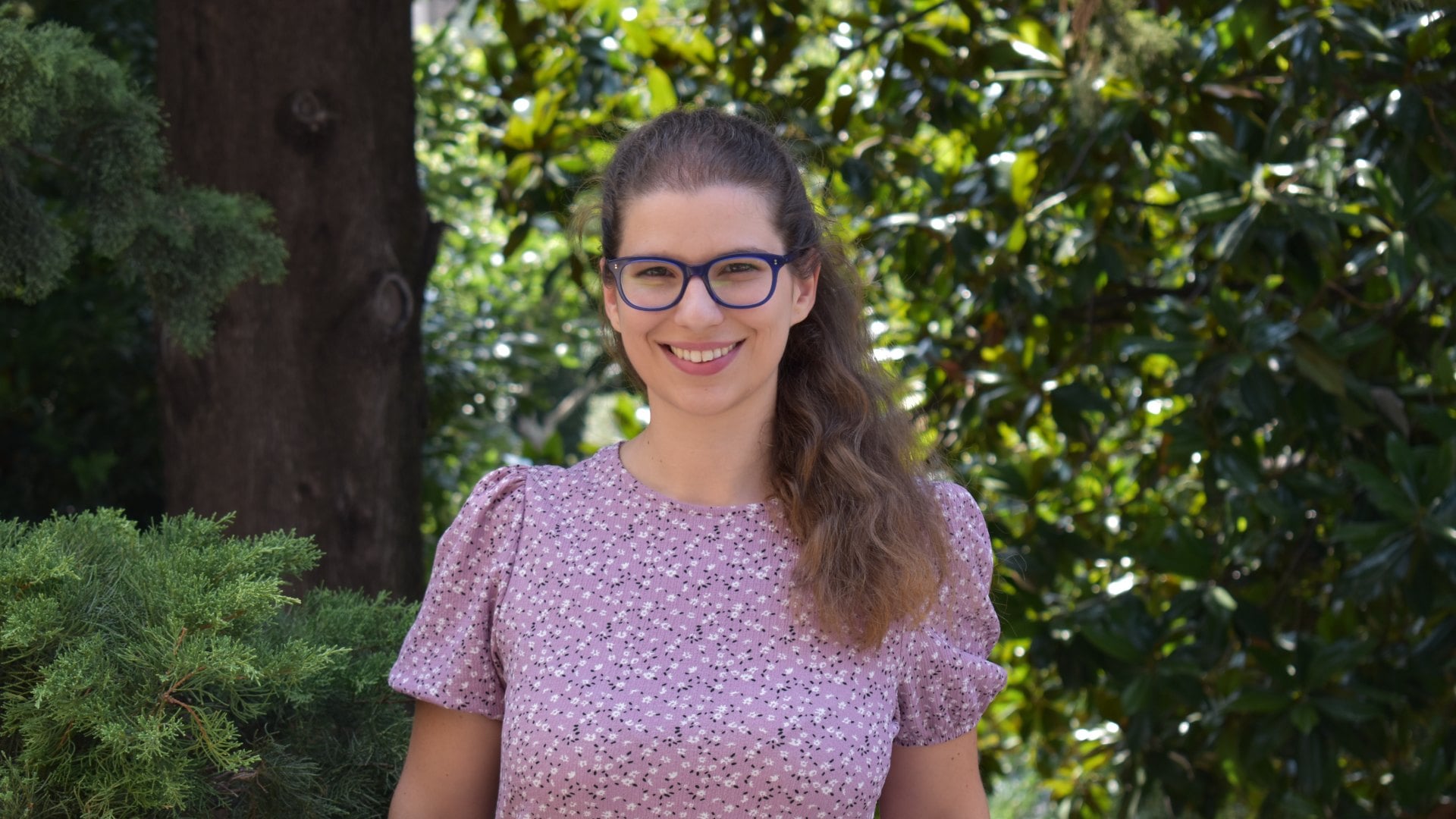 A smiling woman with glasses standing outdoors in front of green trees.