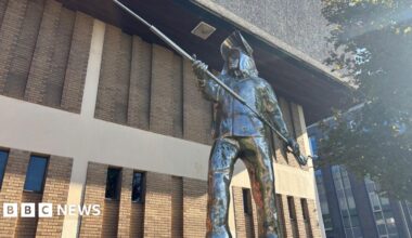 A metal statue showing a life-sized man dressed in steelworkers' uniform and holding a long pole with both hands.