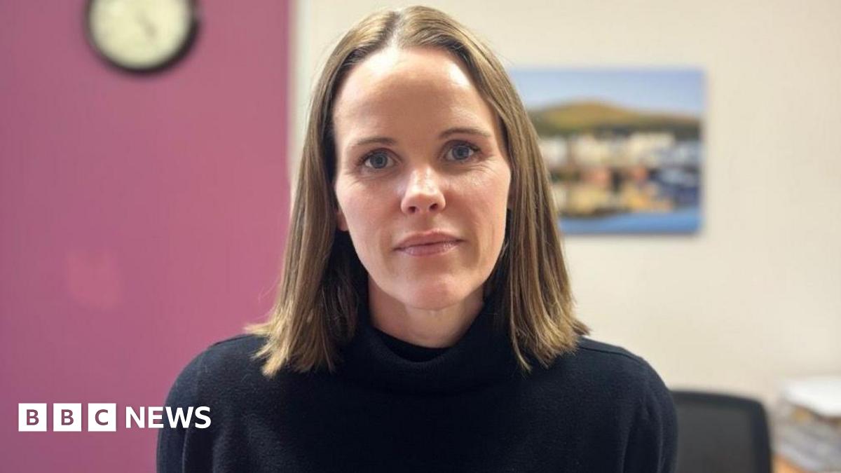 Fiona Hatton has brown shoulder length straight hair and blue eyes. She's wearing a black jumper. She's standing in a room with a purple wall to the left and cream wall with a landscape picture on the wall to the right.