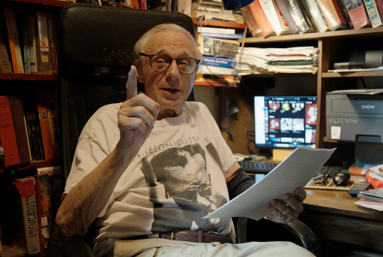 Former Village Voice rock critic Robert Christgau sitting at a desk with a finger raised.