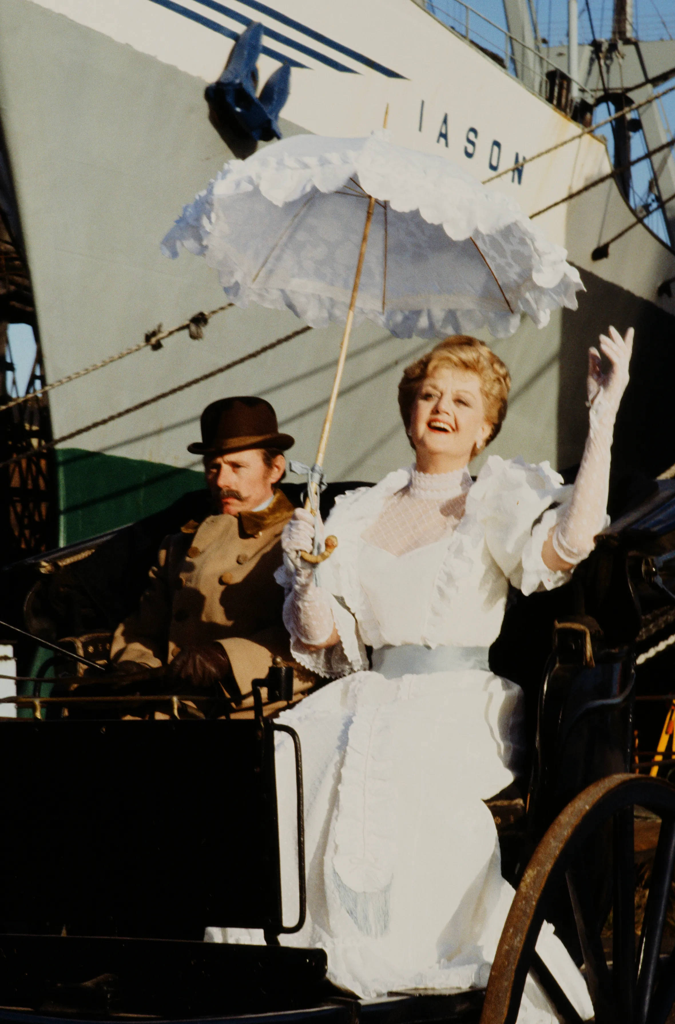 Angela Lansbury as Carole Latimer, holding a white parasol, in a carriage with a mustachioed man, a ship in the background.