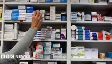 An arm with a grey sleeve is reaching out to a set og shelves packed with a variety of medications
