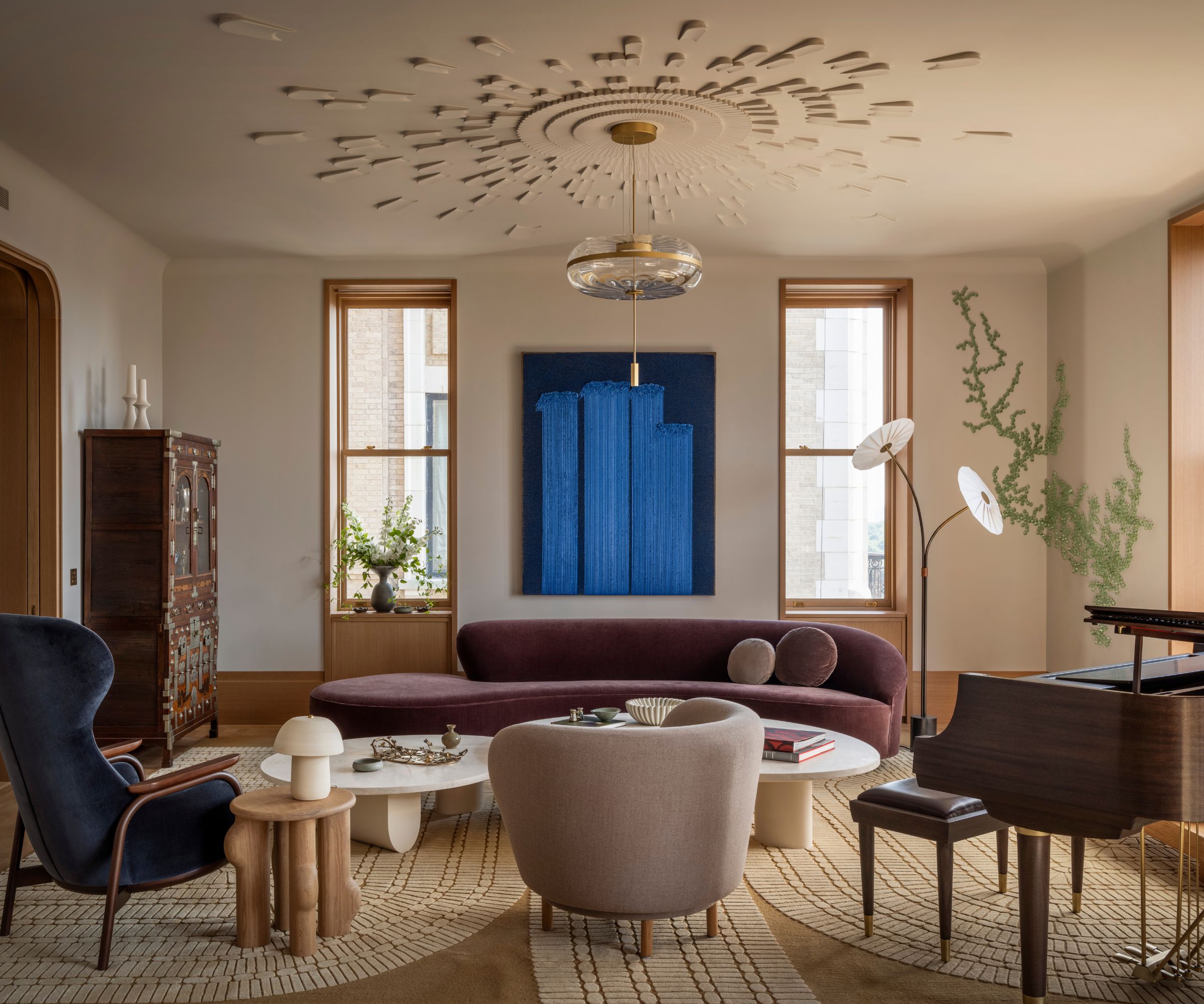 A large living room with off-white walls, a large ceiling rose, two windows with wooden frames, a modern sculptural plum-colored sofa, and a large, patterned neutral rug.