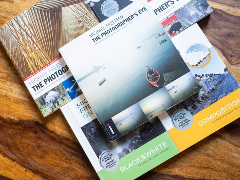Five photography books by Michael Freeman are spread out on a wooden surface, with "The Photographer's Eye" centered on top. The book covers feature various landscape and black-and-white images.