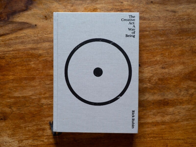 A hardcover book titled "The Creative Act: A Way of Being" by Rick Rubin rests on a wooden surface. The cover is light gray with a simple black circle and dot design in the center.