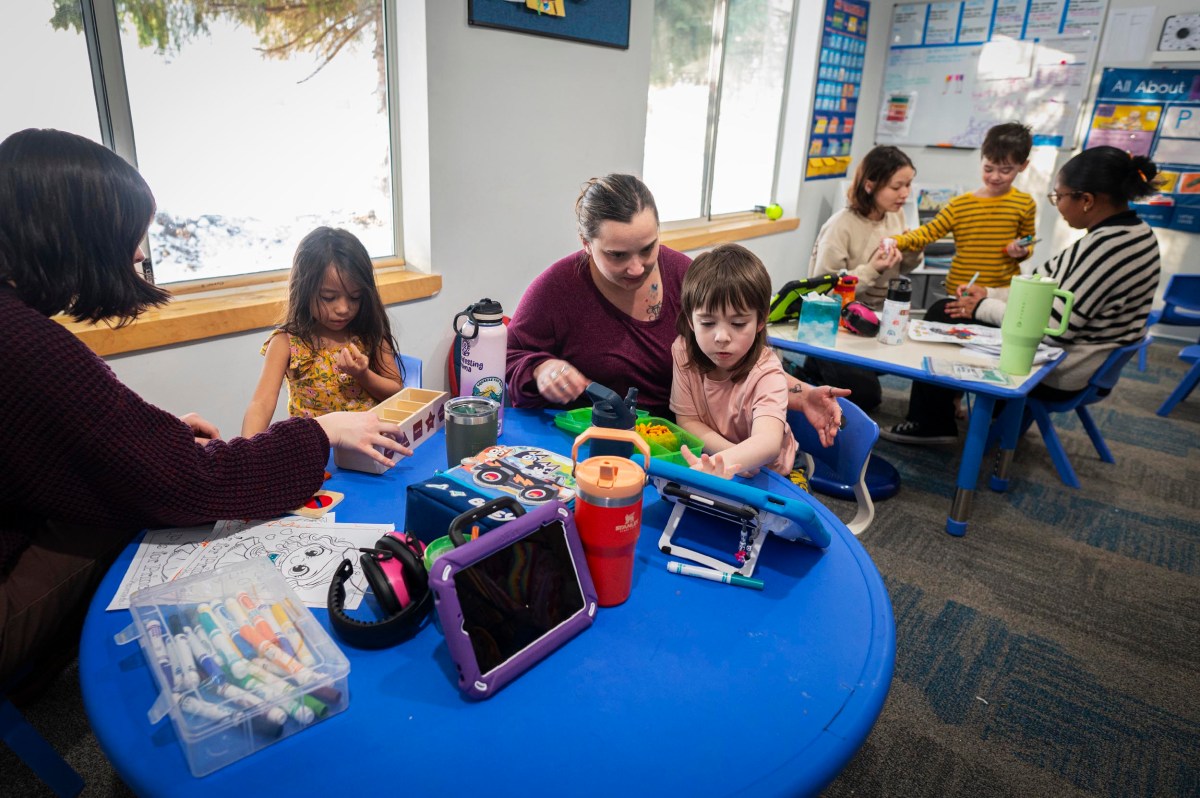 Children and adults sit at blue tables in a classroom, engaging in activities with tablets, coloring supplies, and educational materials.