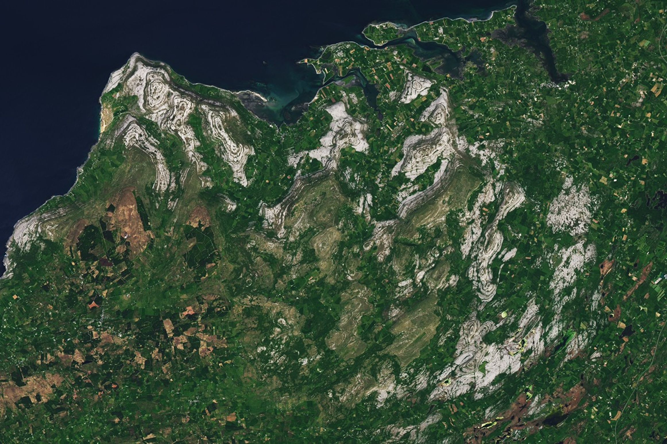 A satellite view of the Burren in western Ireland shows gray limestone hills arranged in curved, concentric bands surrounded by green farmland and small towns.